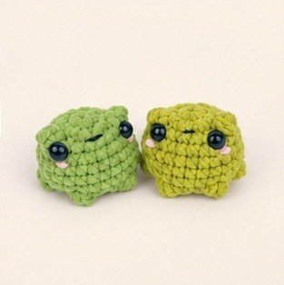 Two tiny green crochet frogs with black eyes and pink cheeks